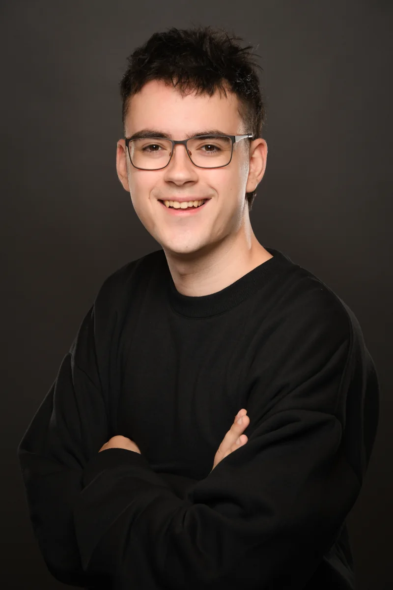 Fruxz lächelt im schwarzen Pullover vor einem dunklen Studiohintergrund.